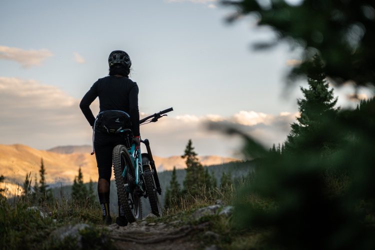 A person standing on a mountain trail, holding a mountain bike, looking out at a scenic landscape with rolling hills and a partly cloudy sky. The figure is wearing a helmet and athletic clothing, surrounded by tall green trees.