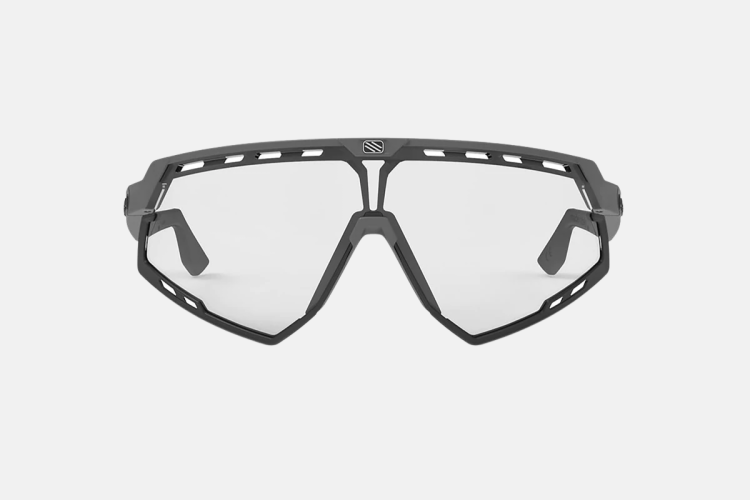 A pair of modern, angular sports sunglasses with a gray frame and clear lenses, featuring cut-out accents along the upper part of the frame for added ventilation and style. The design emphasizes a sporty aesthetic suitable for outdoor activities.