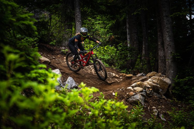 A mountain biker riding a red bike down a dirt trail surrounded by lush green foliage and tall trees. The rider is wearing a helmet and protective gear, navigating a rocky path with visible tire tracks on the ground.