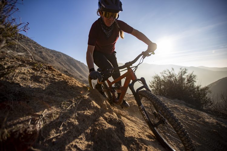 A person riding a mountain bike down a sandy slope during sunset. The rider is wearing a helmet, sunglasses, and gloves, focused on navigating the terrain. The background features a mountainous landscape with soft sunlight filtering through.