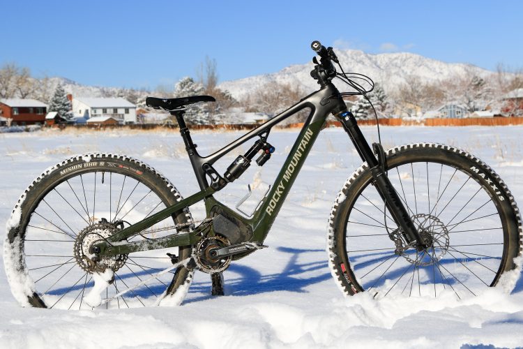 An electric mountain bike stands in a snowy field, showcasing its rugged frame and tires. The bike features a green and black design with visible branding. Snow-covered mountains and trees are in the background under a clear blue sky.