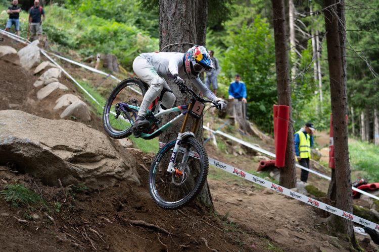 A mountain biker in a white outfit is leaping off a rocky terrain while navigating a downhill course, surrounded by trees and a crowd of spectators. The biker is focused on maintaining balance as they descend, with a visible race number on their bike.