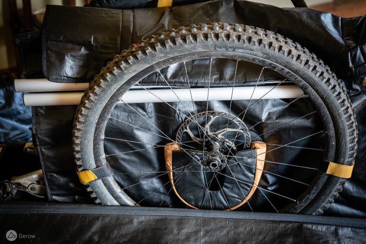 A close-up image of a bicycle wheel partially visible within a black, padded bag. The wheel is mounted with a dirt tread and shows signs of use. There are two white tubes positioned above and a yellow strap securing the wheel. The background consists of additional black fabric, suggesting storage or transportation of cycling equipment.
