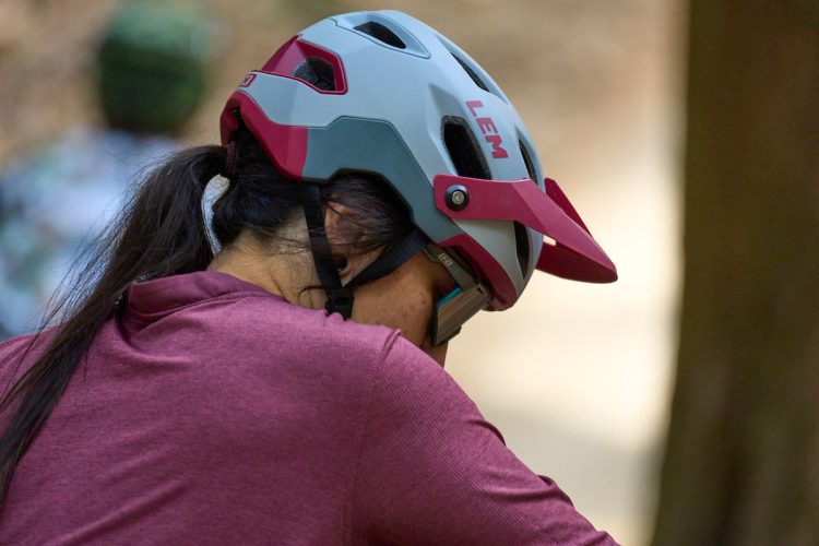 A person wearing a red and gray mountain biking helmet and sunglasses, looking down and resting their arm on their knee. The background is blurred with hints of trees and other bikers.