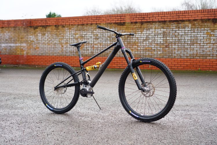 A black mountain bike with distinctive yellow and purple accents, showcasing thick tires and a sturdy frame. The bike features a suspension system and is positioned on a flat, wet surface with a textured brick wall in the background.