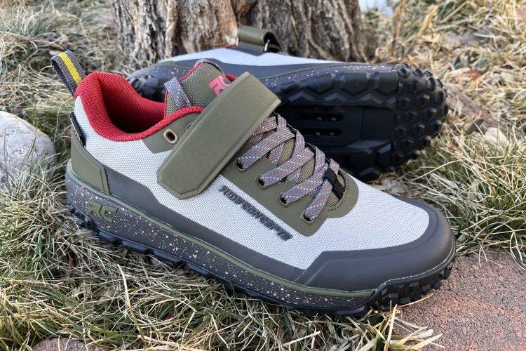 A pair of rugged outdoor shoes featuring a combination of grey, green, and red colors. The shoes have a sturdy design with a textured sole for traction and a reinforced toe cap. They are placed on grass and rocky ground, highlighting their suitability for outdoor activities.