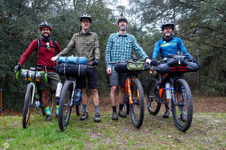 Four cyclists pose together in a wooded area, each standing beside a bicycle equipped for touring. They are wearing helmets and outdoor clothing suitable for biking. Two cyclists have backpacks attached to their bikes, while the other two have various gear and hydration packs. The scene features a mix of greenery and dirt, suggesting an outdoor adventure environment.