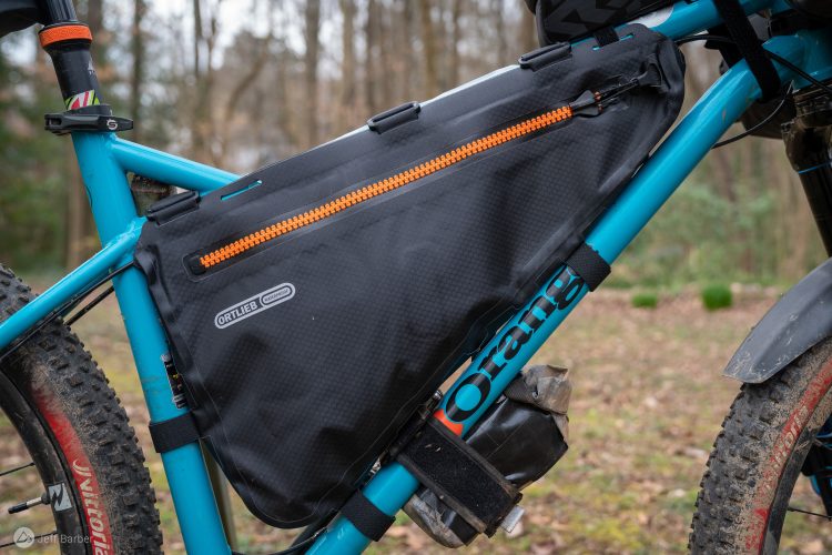 A close-up view of a mountain bike with a black Ortlieb waterproof frame bag attached to the frame, accented by an orange zipper. The bike features a turquoise frame and knobby tires, with a blurred forest background.