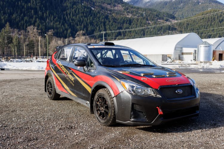 A modified rally car with a black and red exterior featuring the "Crosstrek" logo parked on gravel, with a backdrop of snow and trees in a mountainous area. The vehicle has racing modifications, including a low front bumper and specialized tires, indicating its purpose for off-road competition.