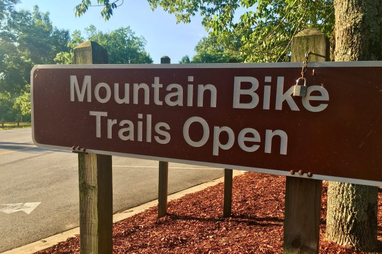 A brown wooden sign displaying the text "Mountain Bike Trails Open," with a padlock hanging from it, surrounded by greenery.
