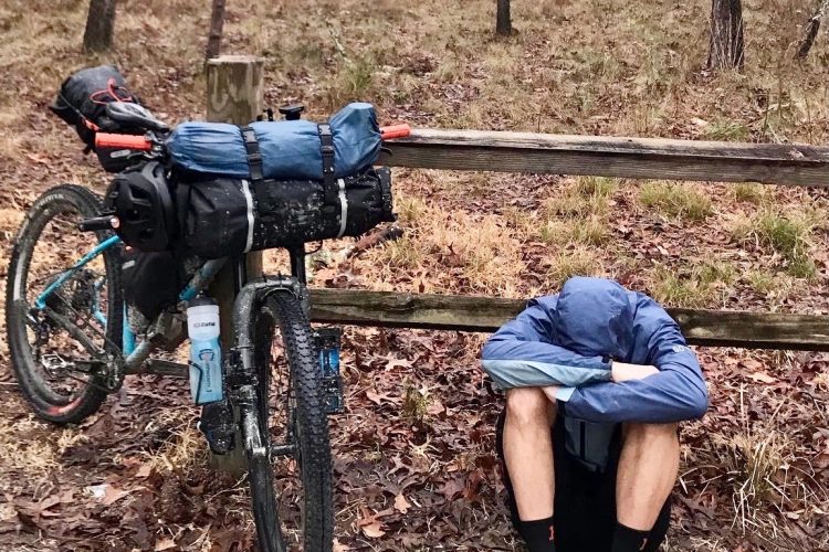 A person sitting on the ground, hunched over with their arms covering their head, next to a bike loaded with camping gear. The scene is set in a rain-drenched forest with fallen leaves and tall trees in the background.