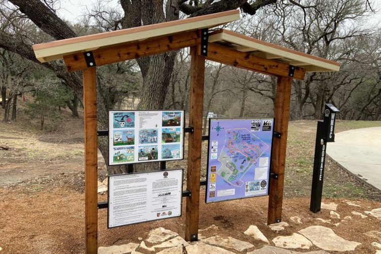 A wooden information kiosk with a slanted roof, displaying a park map and various signs. The kiosk is surrounded by leafless trees and a gravel path. It contains graphics and text providing details about park trails and activities. A "DONATE" sign is visible on the right side of the image.