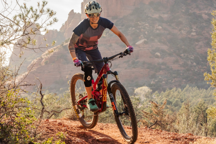 Mountain biker navigating a rocky trail, featuring red rock formations in the background. The cyclist is wearing a helmet, sunglasses, and gloves, and is focused on the path ahead. The scene captures the essence of outdoor adventure and the thrill of mountain biking.