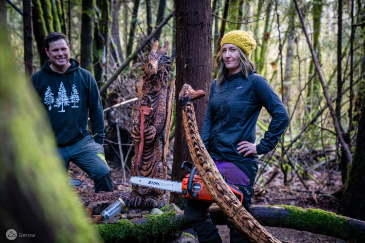 Two people stand in a forest setting, one wearing a black jacket and a yellow knit hat, with a chainsaw at their side. The other person, dressed in a black hoodie with tree graphics, is smiling nearby. They are positioned next to intricately carved wooden sculptures of a dragon and another figure. Surrounding them are tall trees covered in moss.