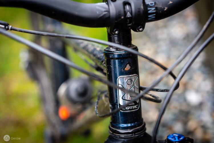 Close-up image of a bicycle's head tube featuring a branded logo. The image captures the handlebars and cables, with a blurred background of greenery and gravel.