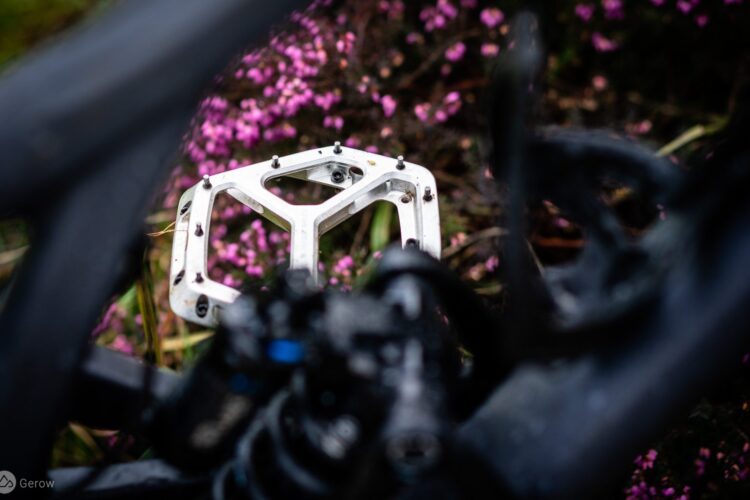 Close-up view of a bicycle pedal, showcasing its metallic structure against a blurred background of purple flowers and green foliage. The pedal features a geometric design with multiple attachment points, reflecting an outdoor setting.