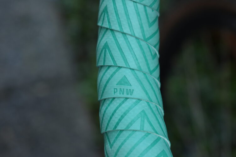 Close-up of a green bicycle handlebar grip with a textured chevron pattern and the letters "PNW" embossed on the surface. The background is out of focus, suggesting a natural setting.