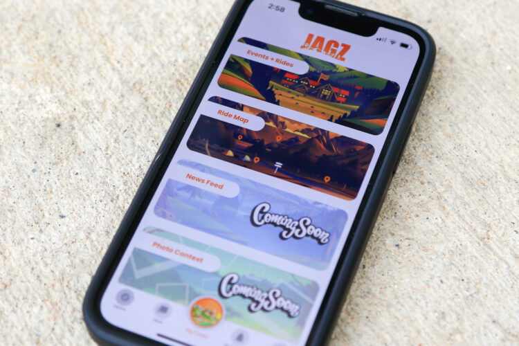A smartphone displaying the user interface of an app called "JAGZ," featuring sections for Events + Rides, Ride Map, News Feed, and Photo Contest, with several items marked as "Coming Soon." The phone is placed on a textured surface.