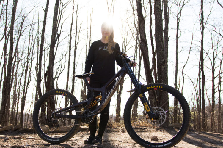 A person stands beside a mountain bike in a forest setting, with sunlight shining behind them, creating a halo effect. The individual is dressed in a black long-sleeve shirt and pants. The bike features large tires and is positioned on a dirt path surrounded by trees with bare branches.