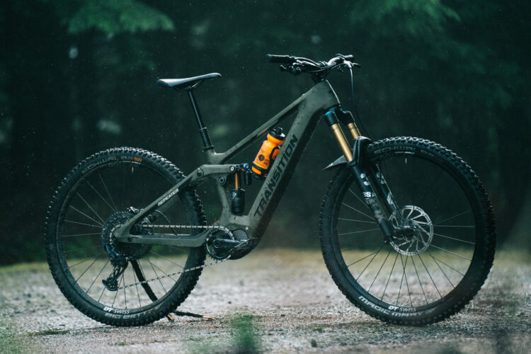 A mountain bike resting on a wet, gravelly surface, surrounded by trees in a rainy setting. The bike features a dark green frame, thick tires, and a visible water bottle mounted on the frame. Raindrops can be seen falling around it, creating a moody atmosphere.