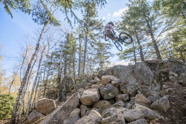 A mountain biker performing a jump over a rocky terrain in a forested area, surrounded by tall trees and a clear blue sky.