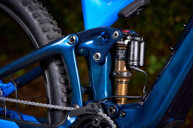 Close-up view of a blue mountain bike frame showcasing the rear suspension system, including a shock absorber and chainstay elements. The background features a blurred natural setting, emphasizing the bike's design and engineering details.