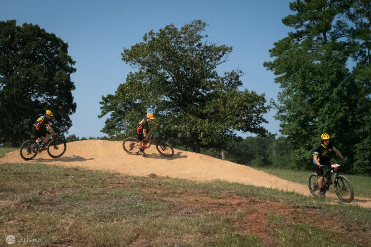 Three young mountain bikers wearing yellow helmets ride along a dirt trail with a mound in a wooded area. The scene captures their action against a backdrop of trees and a clear blue sky.