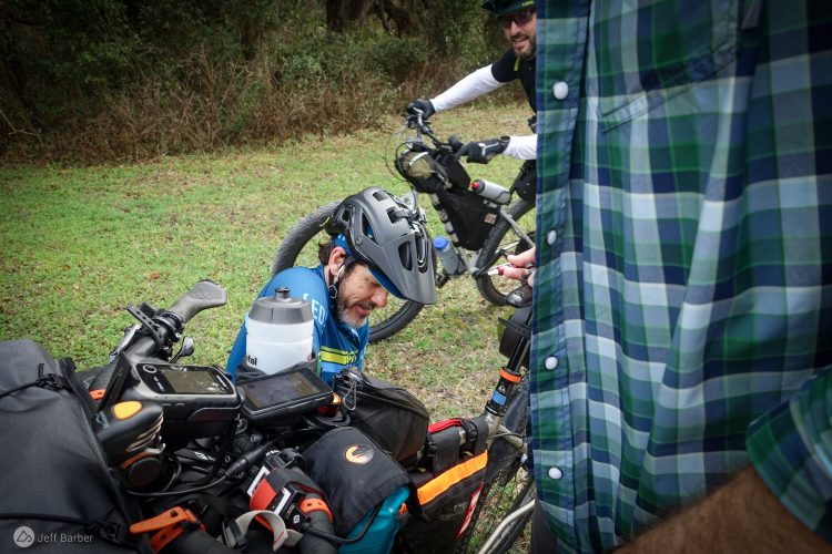 A group of cyclists taking a break outdoors, with one cyclist on the ground inspecting his bike gear. He is wearing a blue cycling jersey and a helmet, while another cyclist stands nearby, partially obscured, wearing a plaid shirt. In the foreground, various cycling equipment and tools are visible on the bikes. The scene is set in a grassy area with trees in the background.