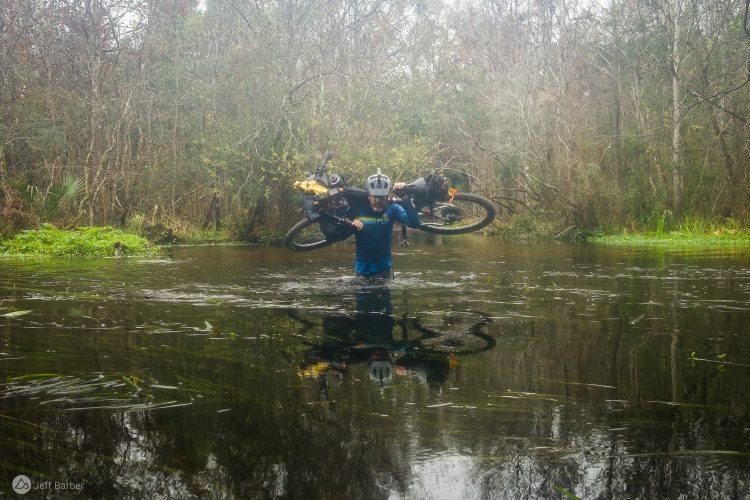 A person wades through water while carrying a mountain bike over their shoulders. The surrounding environment features dense trees and lush vegetation, reflecting the tranquility of a forested area. The water is calm, with patches of floating plants visible. The individual is wearing a blue shirt and a helmet, emphasizing an adventurous spirit in a natural setting.