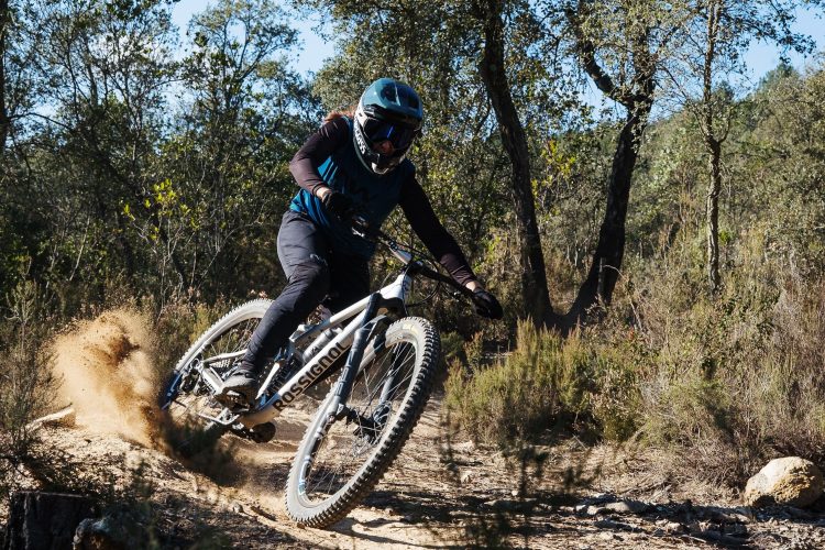 A mountain biker navigating a dirt trail in a forested area, kicking up dust as they make a sharp turn. The cyclist is wearing a helmet and protective gear, showcasing an adventurous outdoor activity.