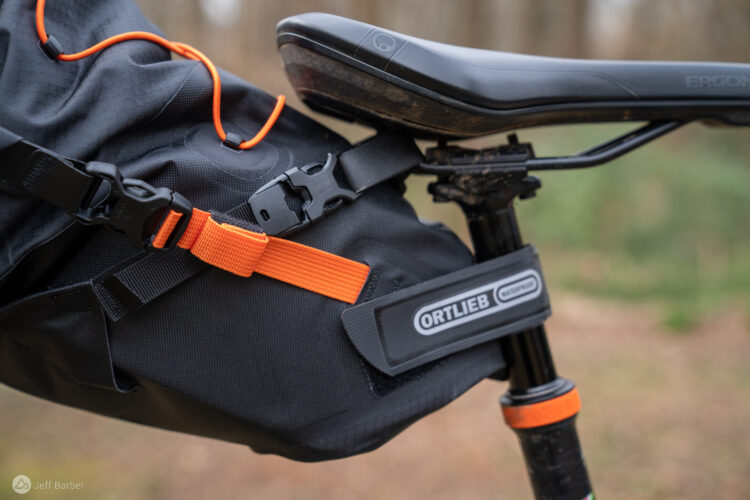 A close-up image of a bike seat and a attached bike bag. The bag is secured with an orange strap and buckle, with the Ortlieb logo visible on the side. The background features blurred natural scenery, indicating an outdoor setting.