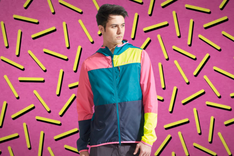 A young man wearing a colorful athletic jacket with pink, green, and navy blue panels stands against a bright pink background adorned with black and yellow geometric shapes.