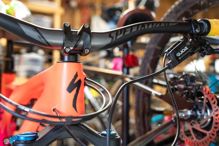 A close-up view of the handlebars and headset of a bright orange mountain bike, featuring a black stem with multiple screws and cables. The bike is set against a cluttered background that includes tools and bike parts, emphasizing a workshop environment.