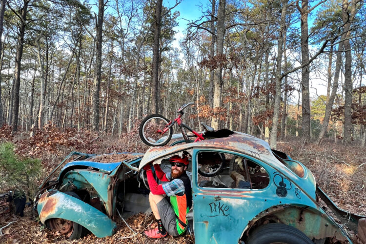 A man wearing a helmet and colorful clothing sits in the driver's seat of a rusted, abandoned car in a forested area. A bicycle rests on top of the car. The scene is surrounded by trees with autumn leaves on the ground under a clear blue sky.