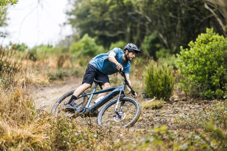 A mountain biker in a blue shirt and black shorts leans into a turn on a gravel trail, surrounded by greenery and tall grasses. Dust and debris fly from the bike's tires, showcasing an action-packed moment in an outdoor setting. The rider is wearing a black helmet and is focused on navigating the challenging terrain.