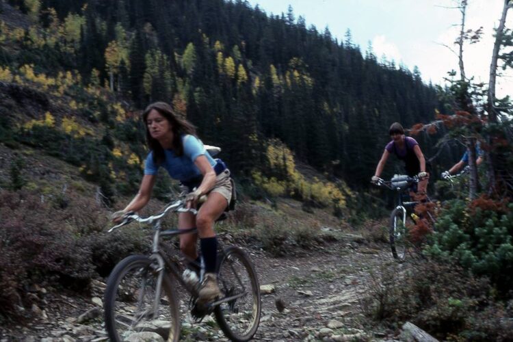 Three people mountain biking on a narrow trail surrounded by trees and autumn foliage. One cyclist, wearing a blue shirt and shorts, is in the foreground, focused on navigating the terrain. Two other cyclists are visible in the background, all riding their bikes on a rugged, natural path.