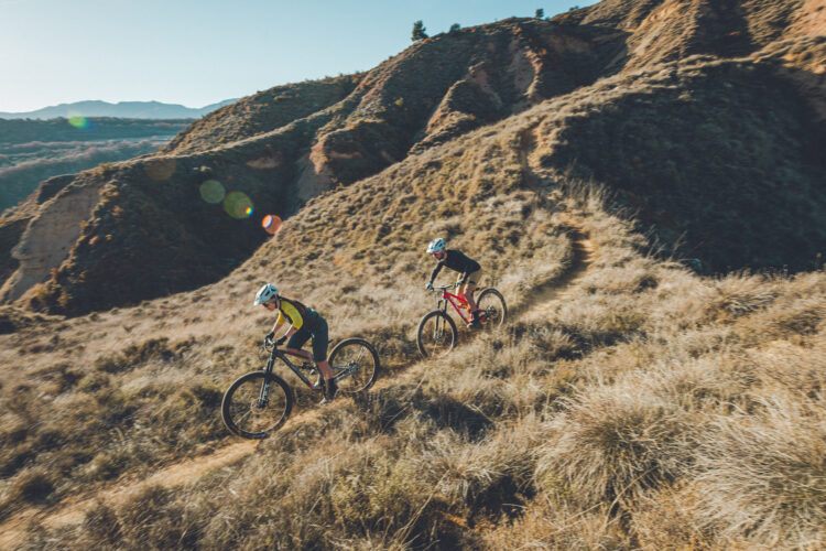 Two mountain bikers navigate a winding dirt trail through a hilly terrain covered with dry grass, against a backdrop of distant mountains and clear blue skies. One rider is dressed in a black shirt and red shorts, while the other is wearing a yellow and green jersey. The scene captures the excitement of outdoor cycling in a natural landscape.