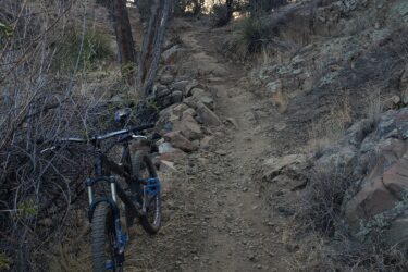 Mountain bike parked along a rocky trail surrounded by sparse vegetation and trees, leading into a wooded area. The afternoon sky is visible in the background, hinting at a serene outdoor setting. Fort Bayard Trail System mountain bike trail.