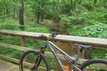 A mountain bike stands parked on a wooden bridge overlooking a small creek, surrounded by lush green trees and foliage in a serene forest setting. The scene captures the tranquility of nature and the excitement of outdoor biking adventures. Tyler State Park mountain bike trail.
