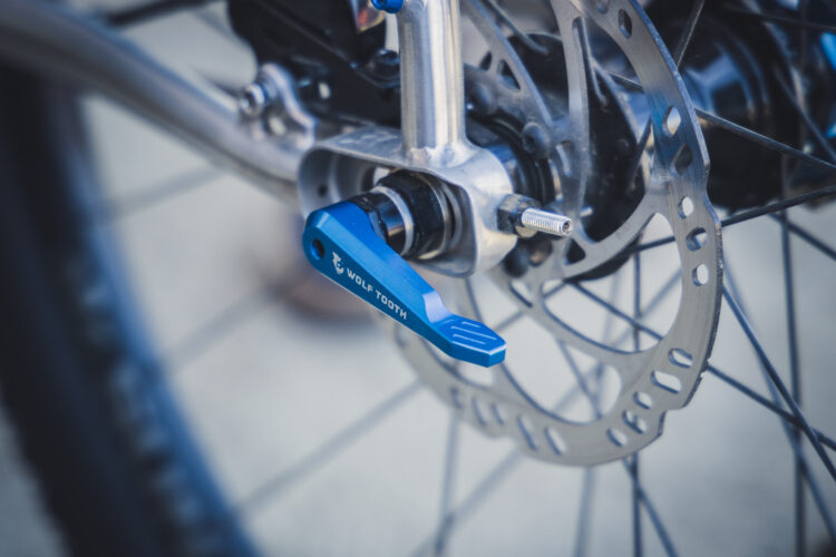 Close-up of a bicycle's rear axle featuring a blue quick release lever by Wolf Tooth, with a disc brake and spokes partially visible in the background.
