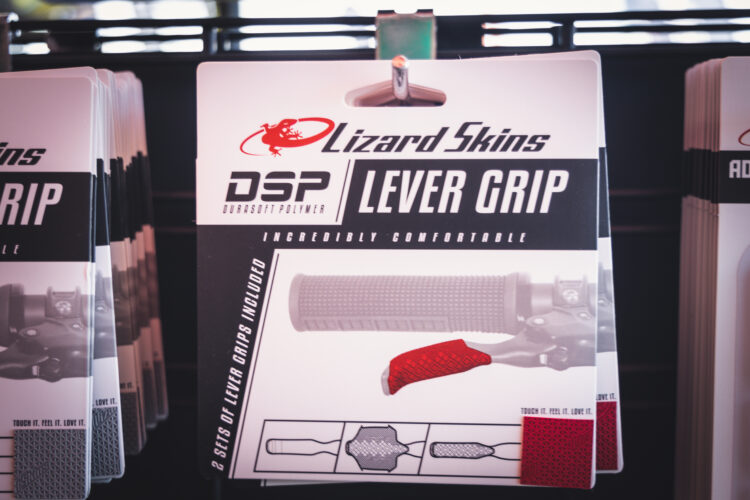A close-up view of packaging for Lizard Skins DSP Lever Grip, featuring images and text that highlight the grip's comfort and design. The packaging includes illustrations of the grip and two color options, red and gray, with captions that say "Incredibly Comfortable" and "2 Sets of Lever Grips Included." The product is displayed in a retail setting.