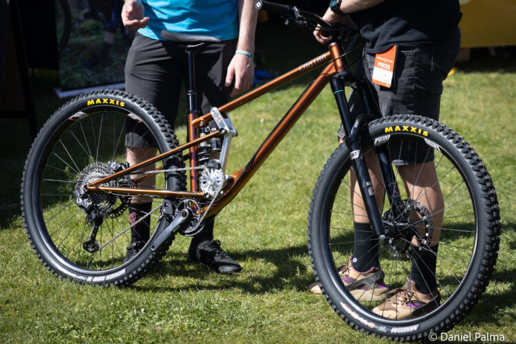 A close-up of a mountain bicycle with a shiny bronze frame, showcased in a grassy outdoor setting. Two individuals are engaged in conversation near the bike, one holding the handlebars. The bicycle features Maxxis tires and a detailed gear system, highlighting its design for performance.