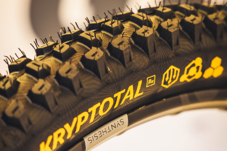 Close-up image of a mountain bike tire featuring the name "KRYPTOTAL" in bold yellow text, showcasing the textured tread pattern designed for traction. The sidewall is labeled with branding details, including the term "SYNTHESIS." The background is softly blurred, emphasizing the tire's design.