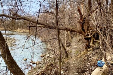 A narrow dirt path bordered by bare trees alongside a river, with scattered rocks and debris along the water's edge. A fallen tree leans over the path, while a piece of concrete and a blue object are visible on the ground to the right. The sky is slightly overcast, creating a muted atmosphere. Etobicoke Creek mountain bike trail.