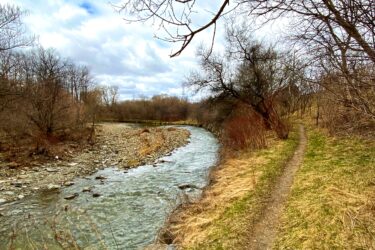 A winding river flows alongside a narrow path bordered by dry grass and sparse trees under a partially cloudy sky, creating a tranquil nature scene. Etobicoke Creek mountain bike trail.