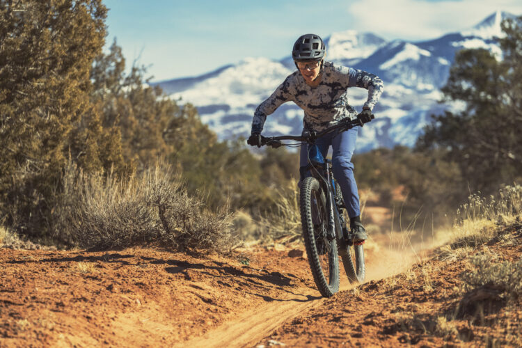 A person riding a mountain bike on a dirt trail surrounded by natural landscape, with a backdrop of snow-capped mountains. The rider is wearing a helmet and is captured in motion, focused and enjoying the outdoor activity. The ground is sandy with patches of grass and shrubbery.