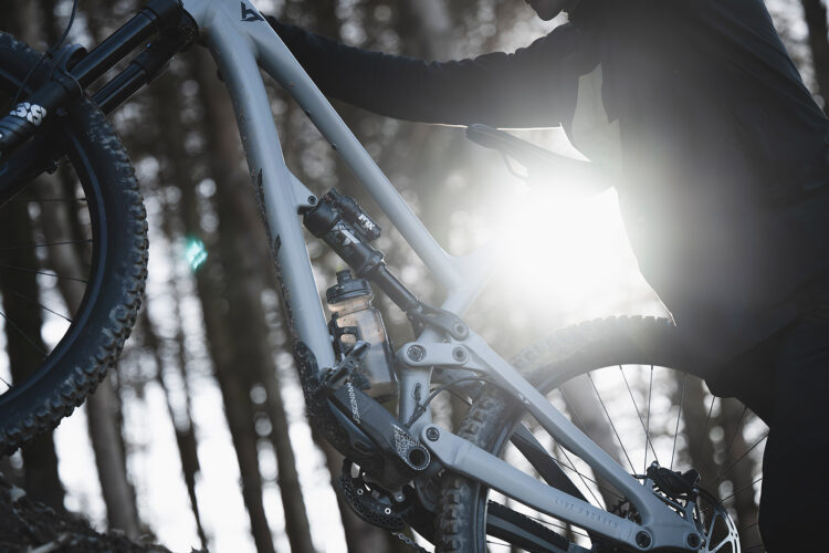 A person in a black jacket is lifting a mountain bike with a grey frame, set against a backdrop of trees. The sun is shining brightly, creating a lens flare effect behind the bike. The scene captures a moment of outdoor adventure and exploration.