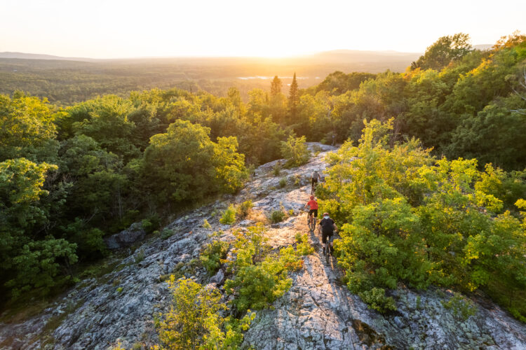 A panoramic view of a rocky landscape at sunset, with four mountain bikers navigating a steep, gravelly trail surrounded by lush greenery. The sun casts a warm glow over the scene, illuminating the trees and hills in the distance.