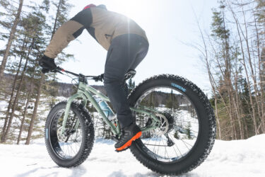 A person riding a fat bike on a snowy trail, surrounded by trees. Sunlight is shining from behind, highlighting the bicycle's large tires and the rider's winter attire. Sentiers Du Moulin mountain bike trail.