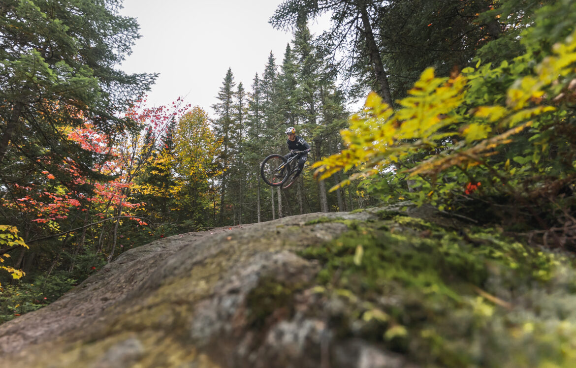 A mountain biker performing a jump over a rock in a forested area, surrounded by trees displaying autumn foliage. The image captures the dynamic movement of the rider in mid-air, with a focus on the natural scenery and vibrant colors of the leaves. Sentiers Du Moulin mountain bike trail.